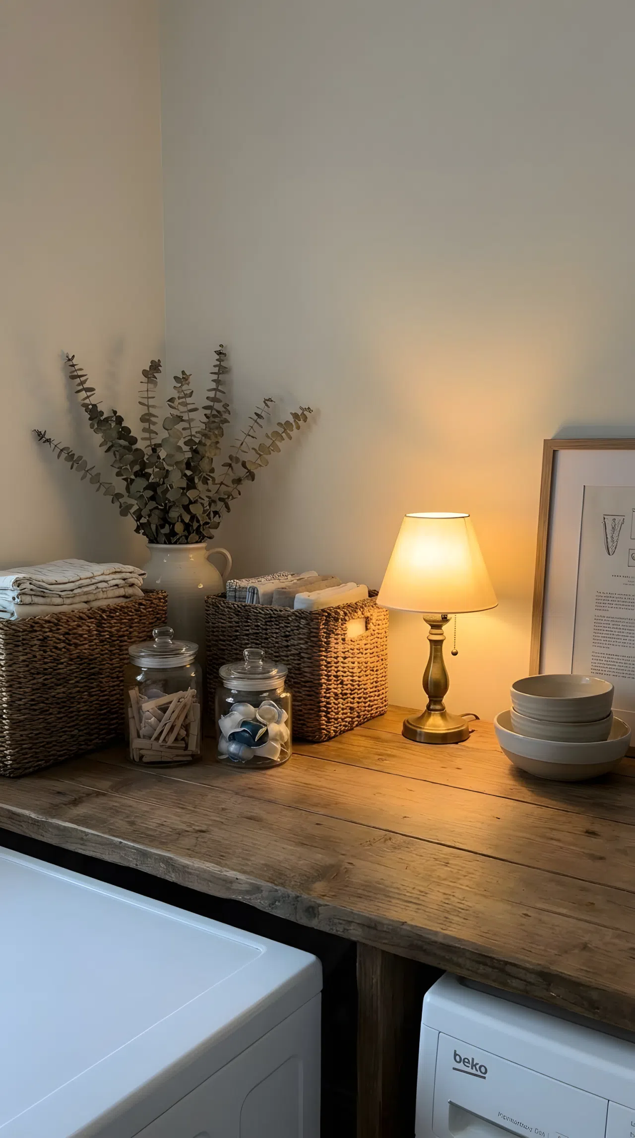 Real-life photography of a small warm toned plug-in lamp placed at counter height beside baskets and jars in a laundry room, casting a low glowing pool of light, counter neatly made and beautifully arranged, organized clean styling, realistic home photography.