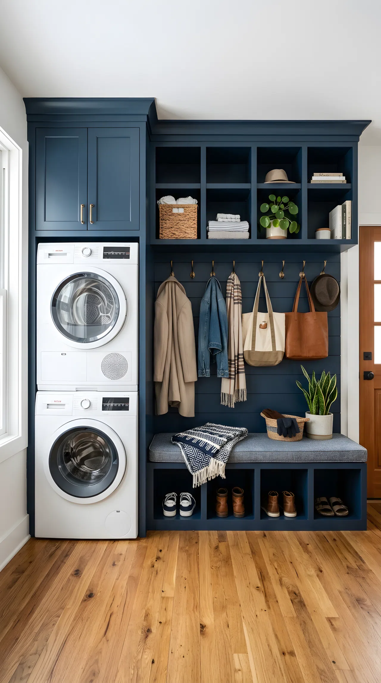 Realistic photograph of one continuous navy blue built-in wrapping a stacked washer and dryer on one end and open cubbies, hooks, and a cushioned bench on the other end, shown together in the same frame. Clean, organized, beautifully arranged space with bright natural light. Authentic real-life stock photo, no illustration or 3D render.