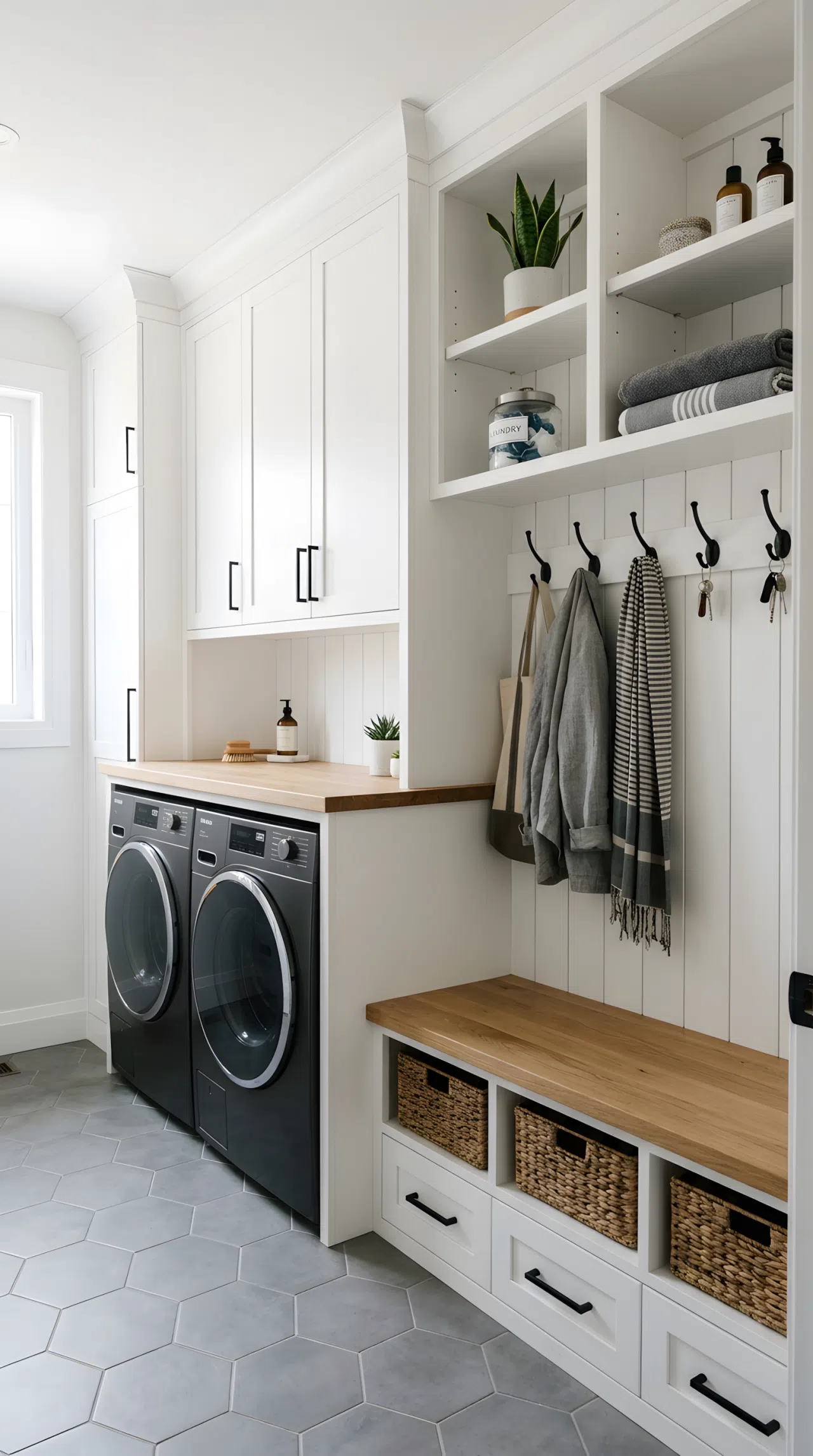 Realistic photograph of one room with crisp white cabinetry covering both a washer-dryer enclosure and a bench-and-hook wall beside it, dark appliance fronts and black hardware repeating across both zones, shown together in a single frame. Clean, organized, beautifully arranged space with bright natural light. Authentic real-life stock photo, no illustration or 3D render.