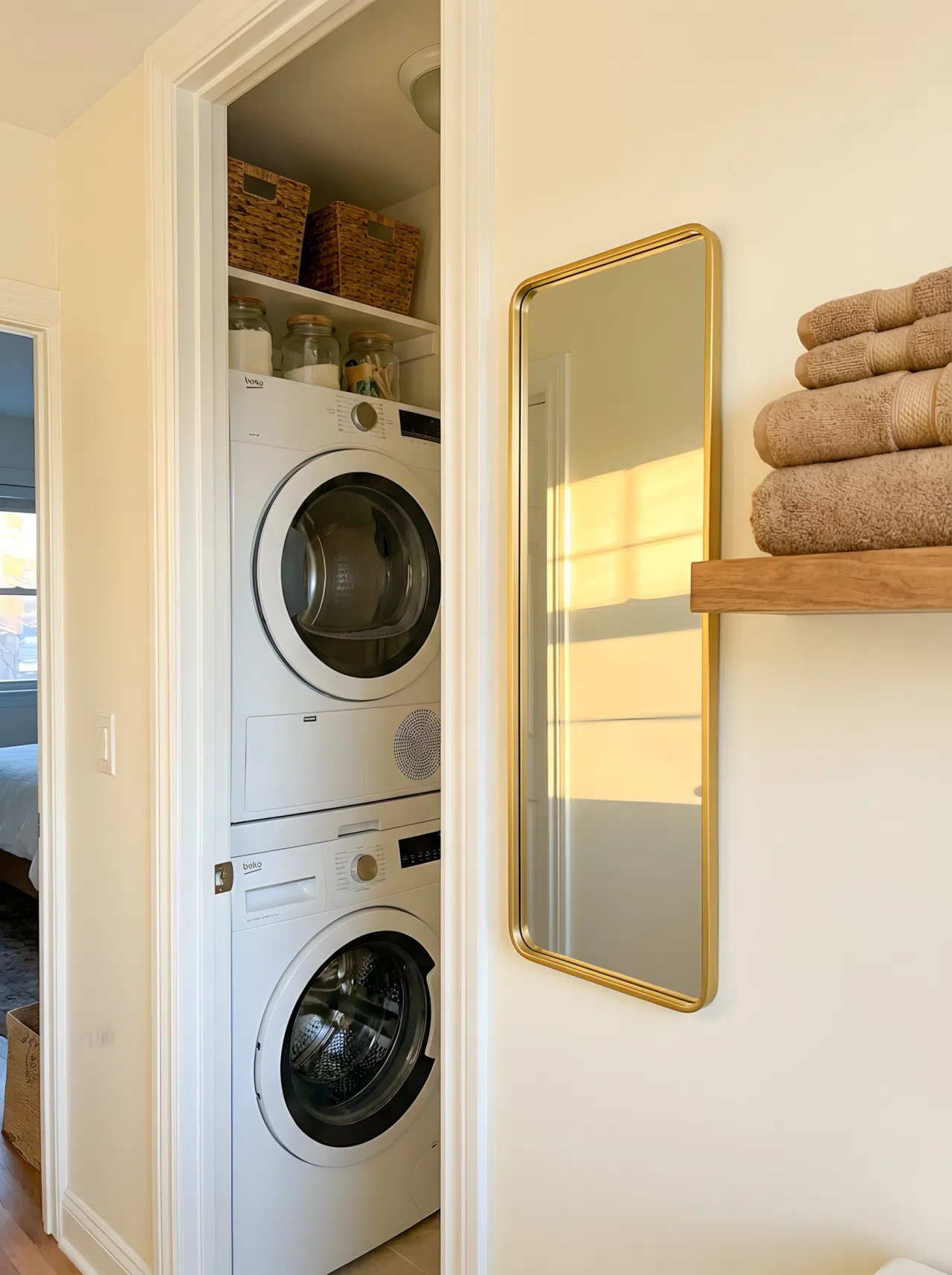 Real-life photograph of a narrow laundry closet with stacked washer and dryer, slim framed mirror mounted on the adjacent wall reflecting warm light, neatly organized shelf with folded towels beside the mirror, bright natural light bouncing off the mirrored surface, clean beautifully arranged space, vibrant tones, eye-level angle, no people