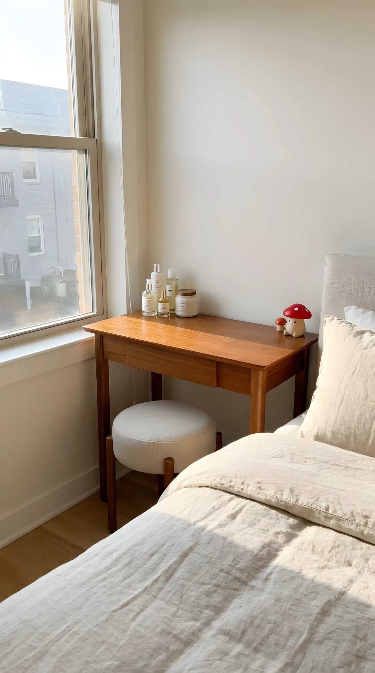 Real-life photo of a slim bedroom vanity placed directly beside the bed, low stool tucked underneath, neatly made surface with a few skincare items, small red mushroom toy figurine on the tabletop, bright natural light, clean and beautifully arranged, vibrant stock photography style.