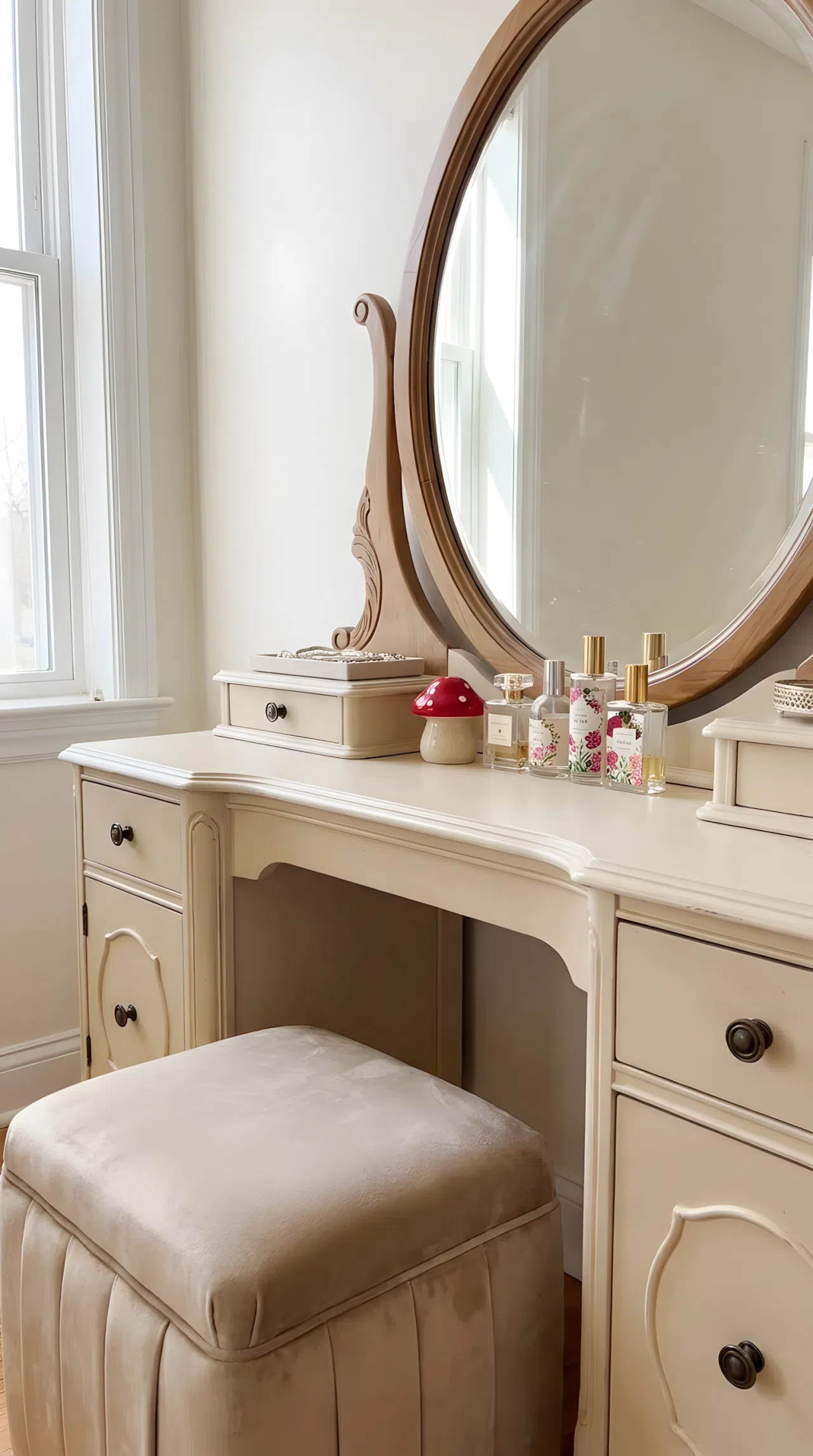 Real-life photo of a vintage bedroom vanity table in soft cream tones (RGB 245, 235, 220) with beige velvet stool, curved mirror, neatly made surface, beautifully arranged perfume bottles, small red mushroom toy figurine placed beside the mirror, bright natural light, clean and organized, elegant vibrant stock photography.