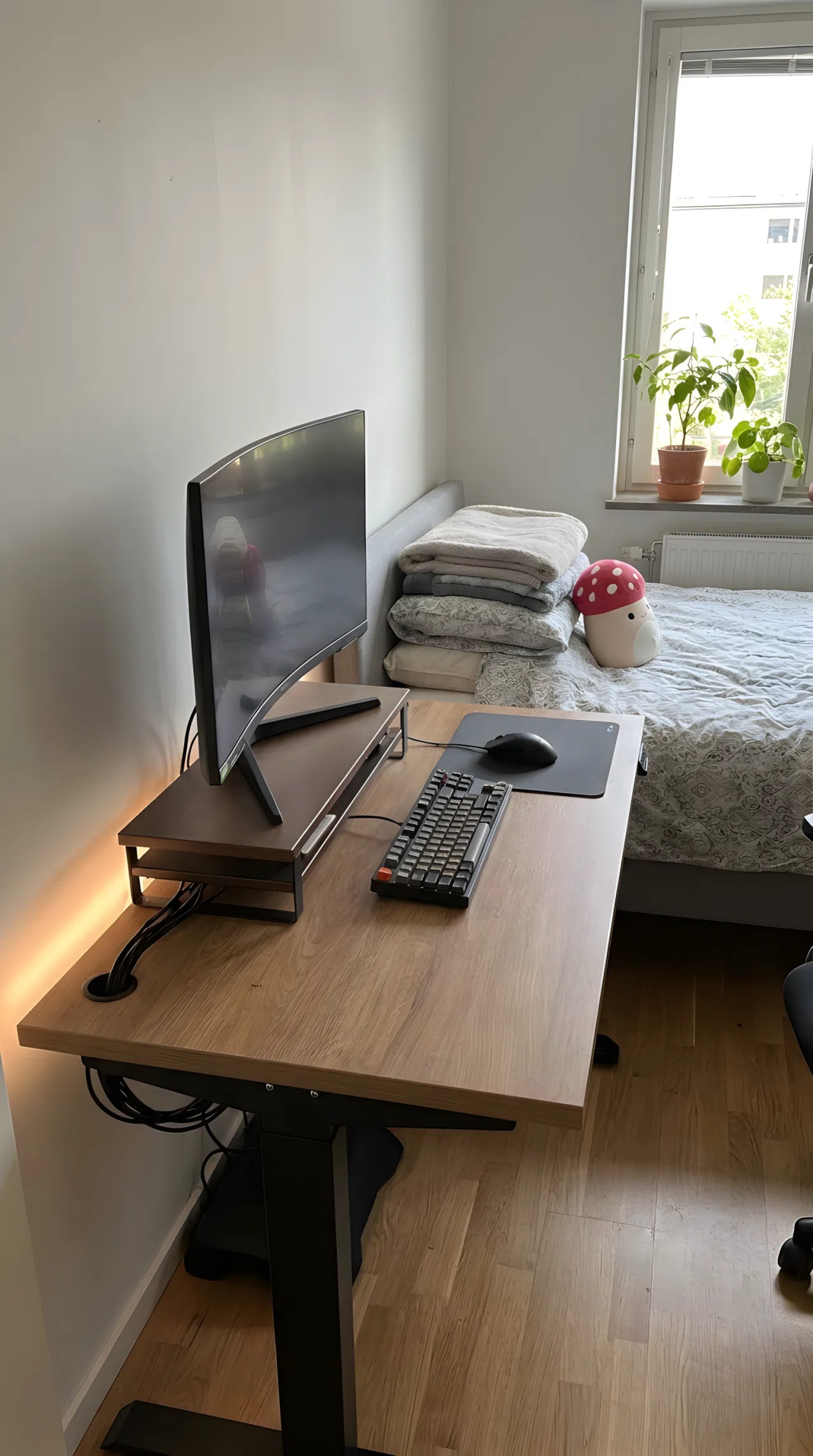 A neatly made bedroom with an organized gaming desk setup featuring a monitor riser and subtle LED strip lighting along the desk edge, small red mushroom-shaped plush toy on a beautifully arranged nearby bed, tidy cable management, bright natural light, clean and organized, real-life lifestyle photography, vibrant colors, no people, no text overlays.