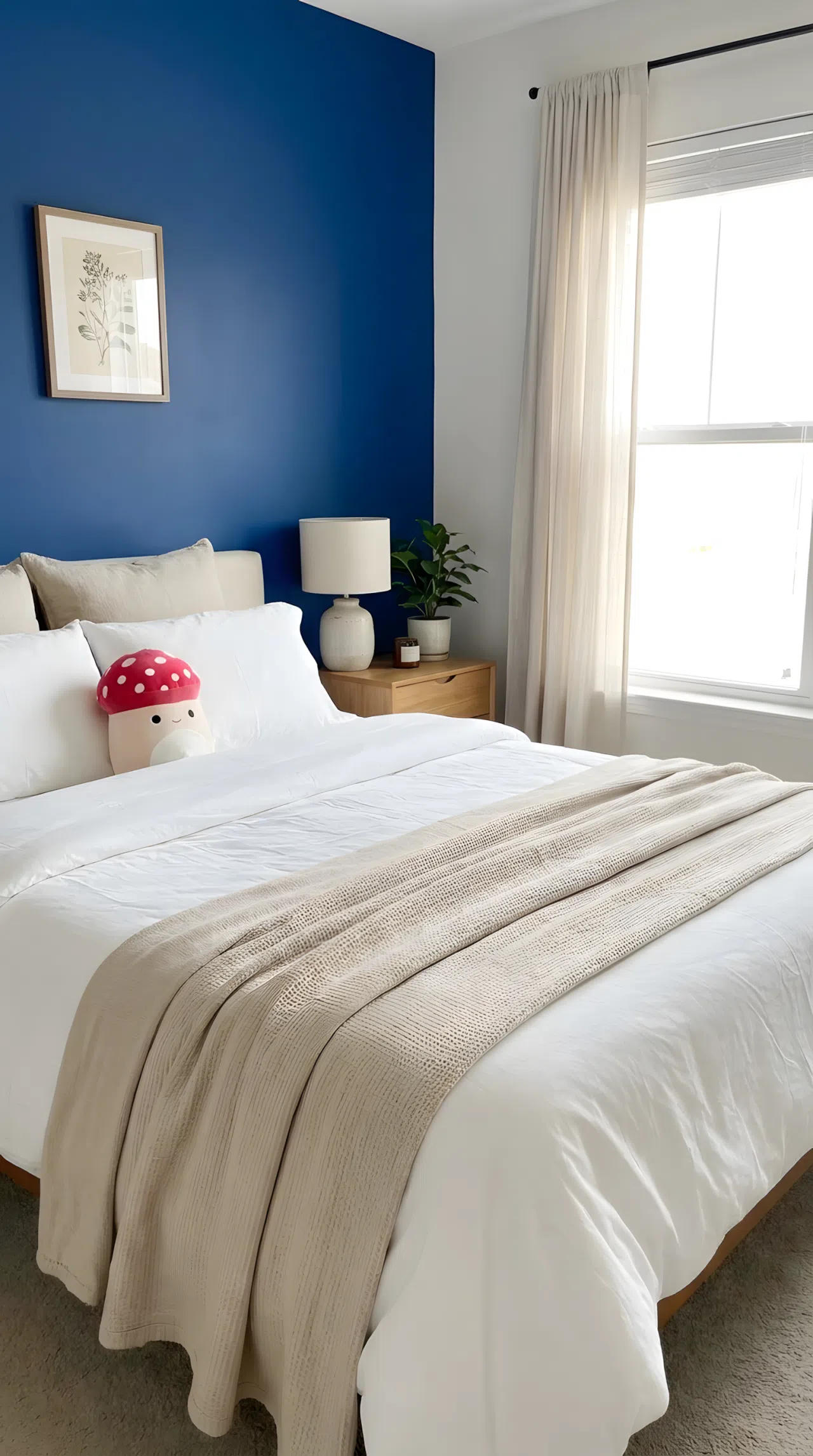 A neatly made bedroom with one accent wall painted deep cobalt blue (RGB 25, 65, 120), a small red mushroom-shaped plush toy sitting on a beautifully arranged white bed, neutral beige bedding, minimal furniture, bright natural light, clean and organized, real-life lifestyle photography, vibrant colors, no people, no text overlays.
