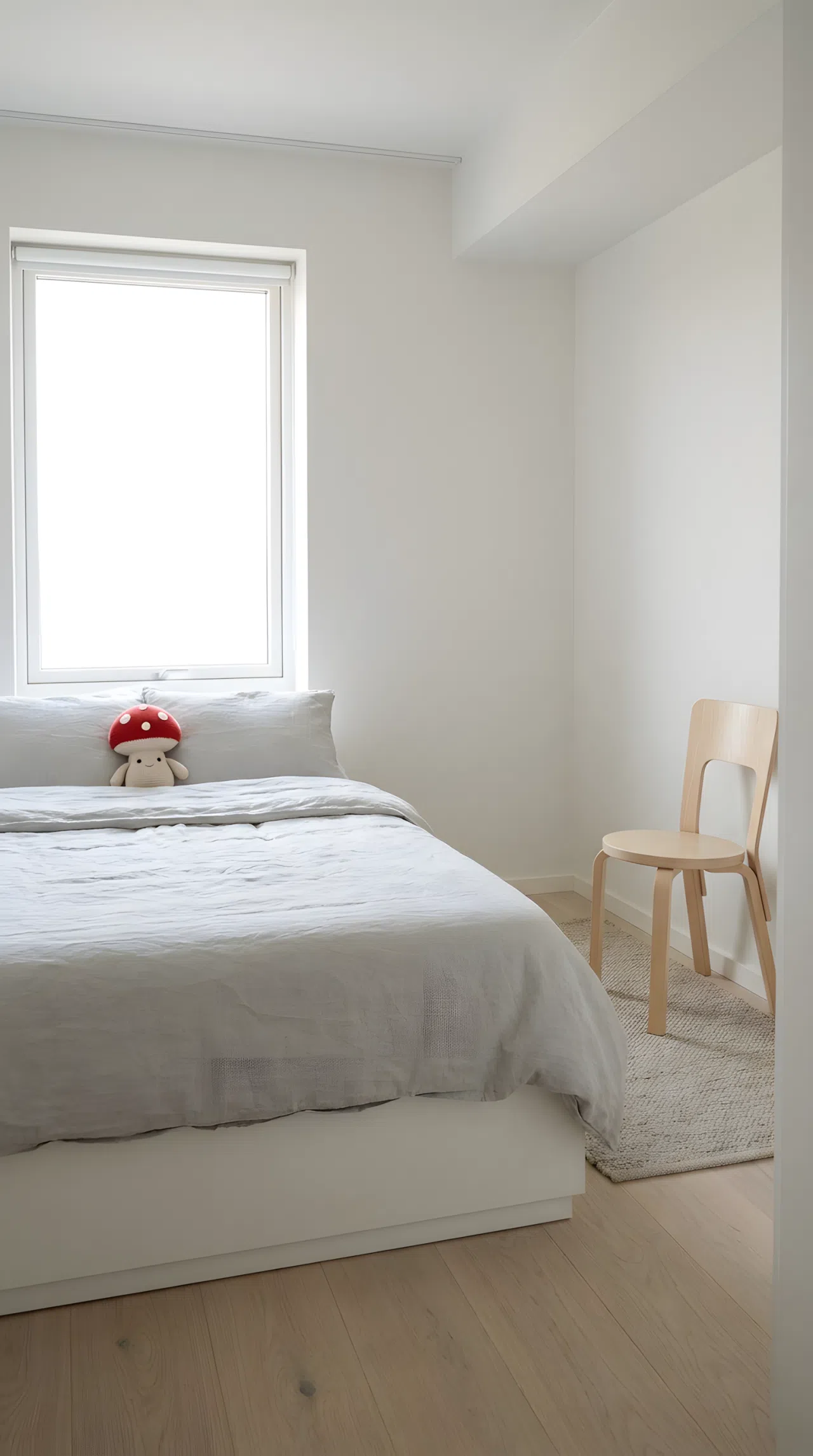 Minimalist Scandinavian bedroom with a neatly made bed, single wooden chair, small red mushroom-shaped plush toy on the pillow, open uncluttered floor space, organized and clean styling, bright natural light through a large window, real-life photography, vibrant soft tones
