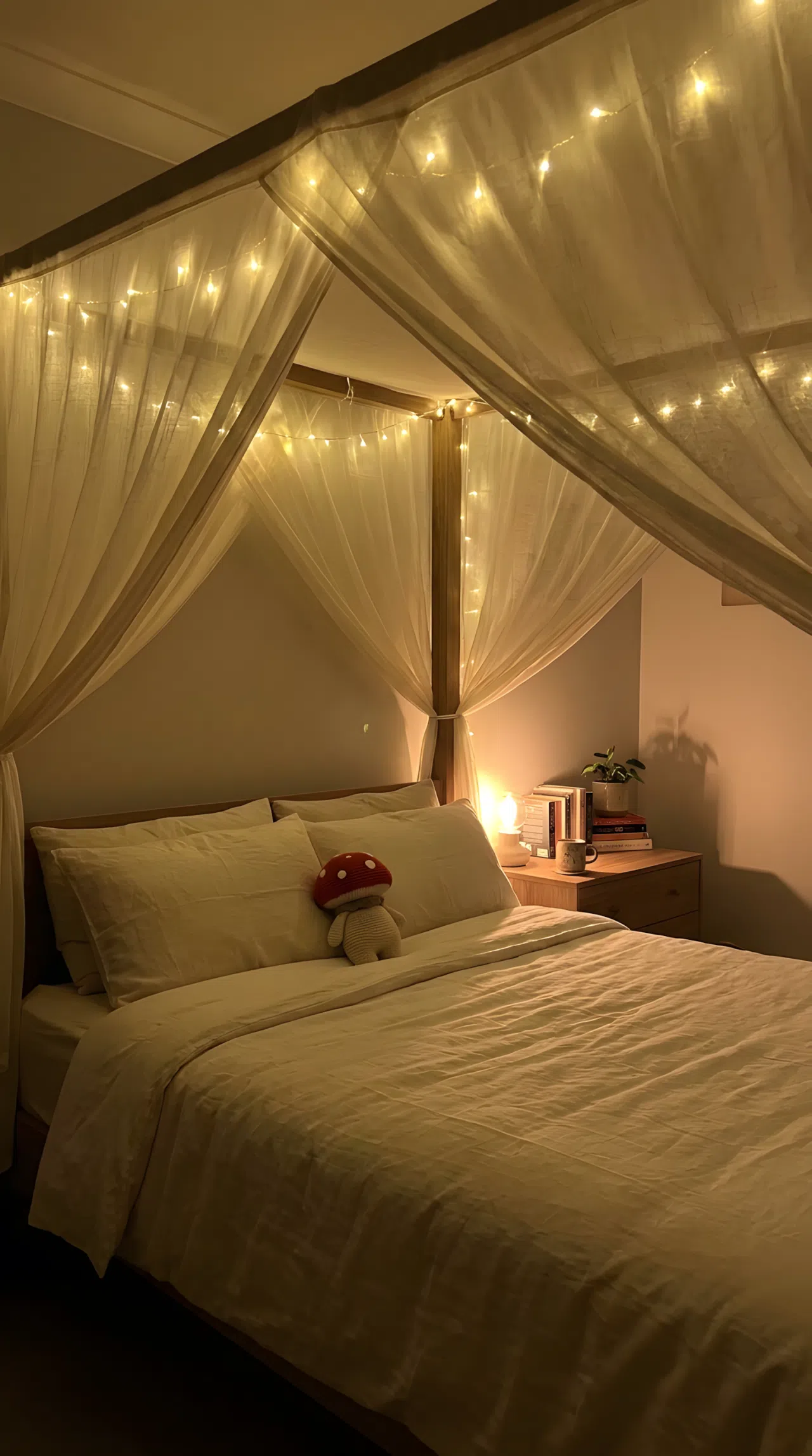 Bed with a canopy of warm fairy lights strung overhead casting a soft downward glow, neatly made bedding, small red mushroom plush toy on the pillow, organized bedside styling, dim ambient lighting, real-life photography, vibrant warm glow, no harsh overhead light