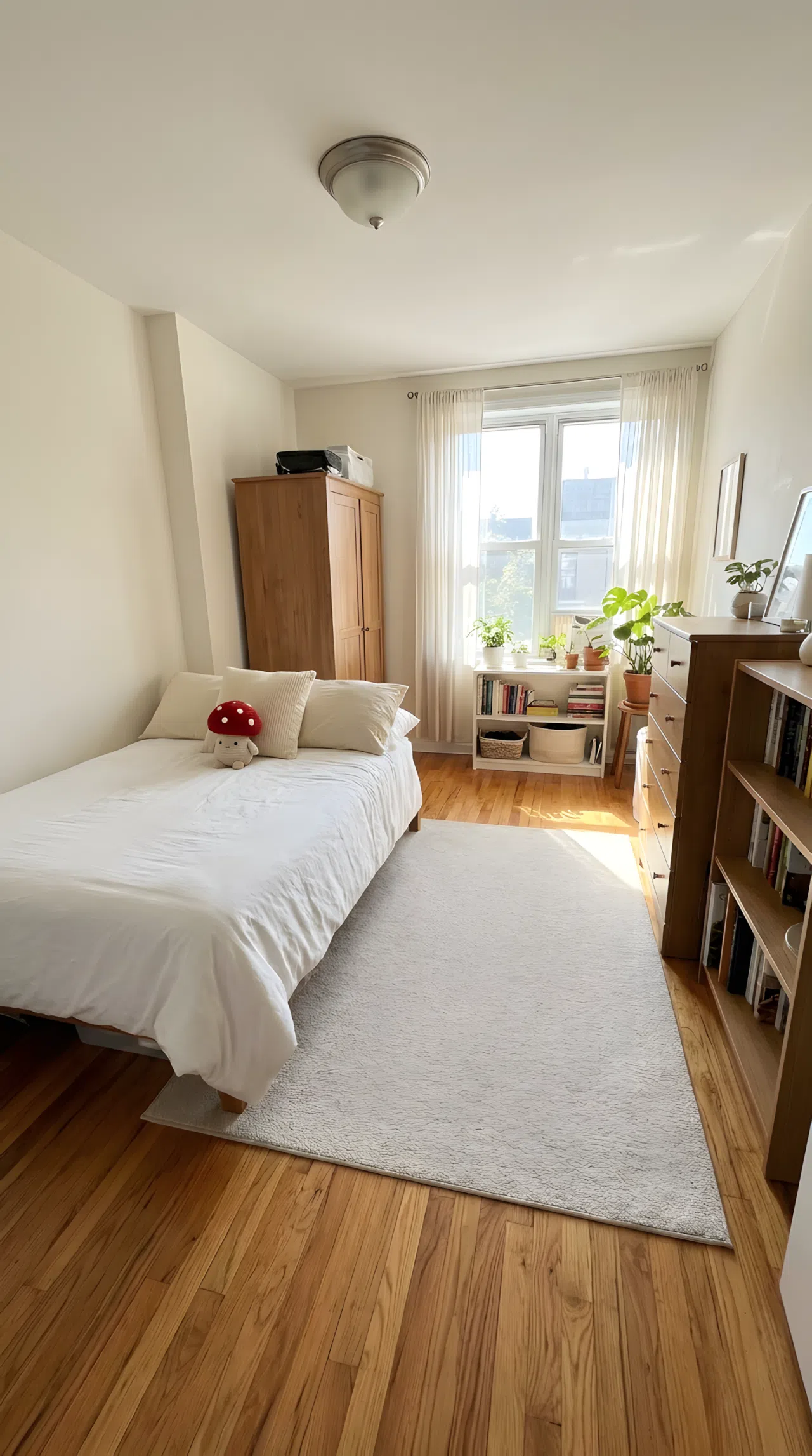 Bedroom with furniture arranged along the walls leaving an open walking path, neatly made bed with small red mushroom-shaped plush toy on the pillow, organized and clutter-free floor, bright natural light, real-life photography, vibrant clean composition, functional layout