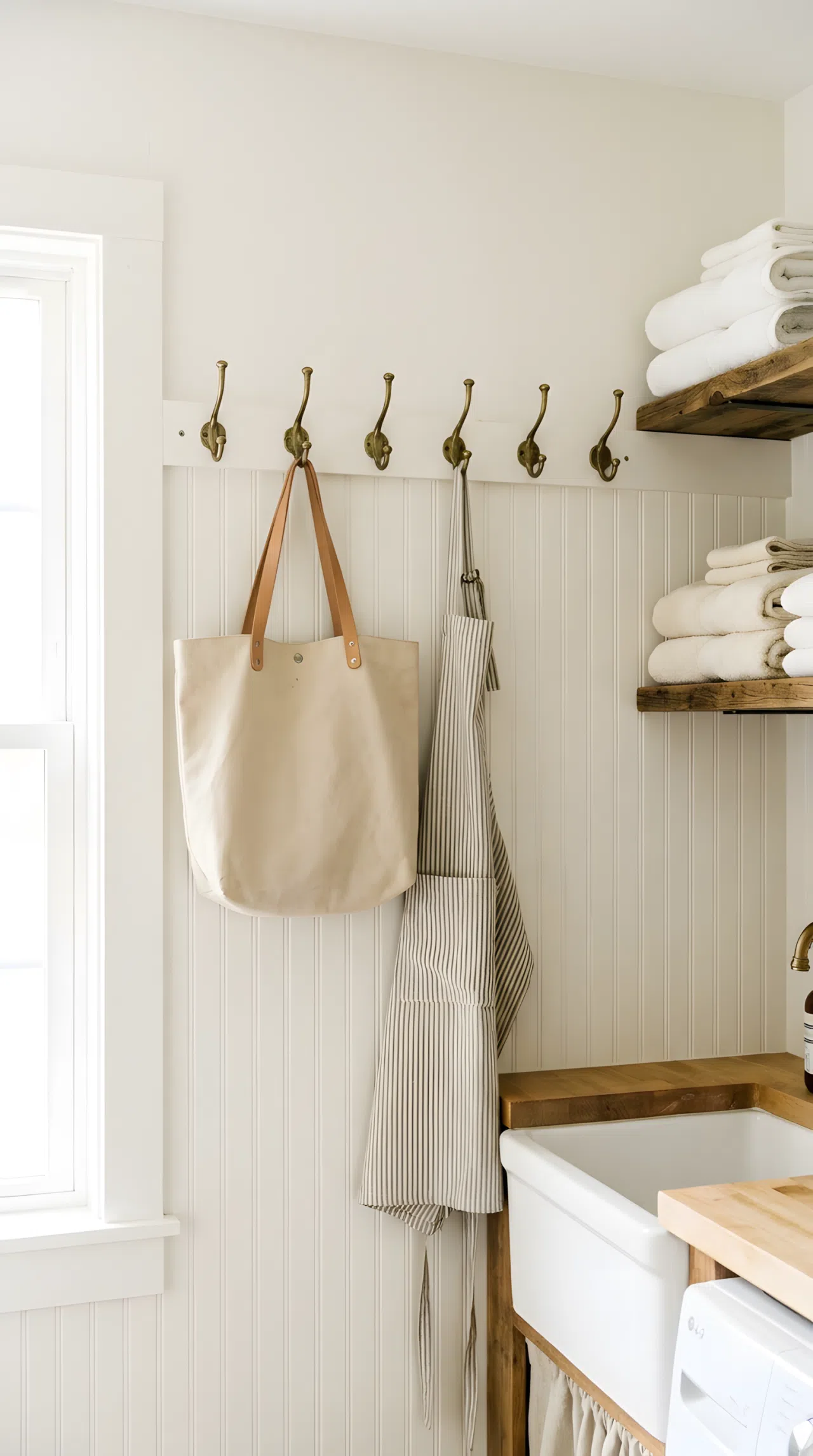 A neatly made farmhouse laundry room wall with a row of antique brass hooks holding a canvas tote bag and apron beautifully arranged, folded towels organized on a nearby shelf, bright natural light filling the space, clean vibrant real-life photography, no people, no text.