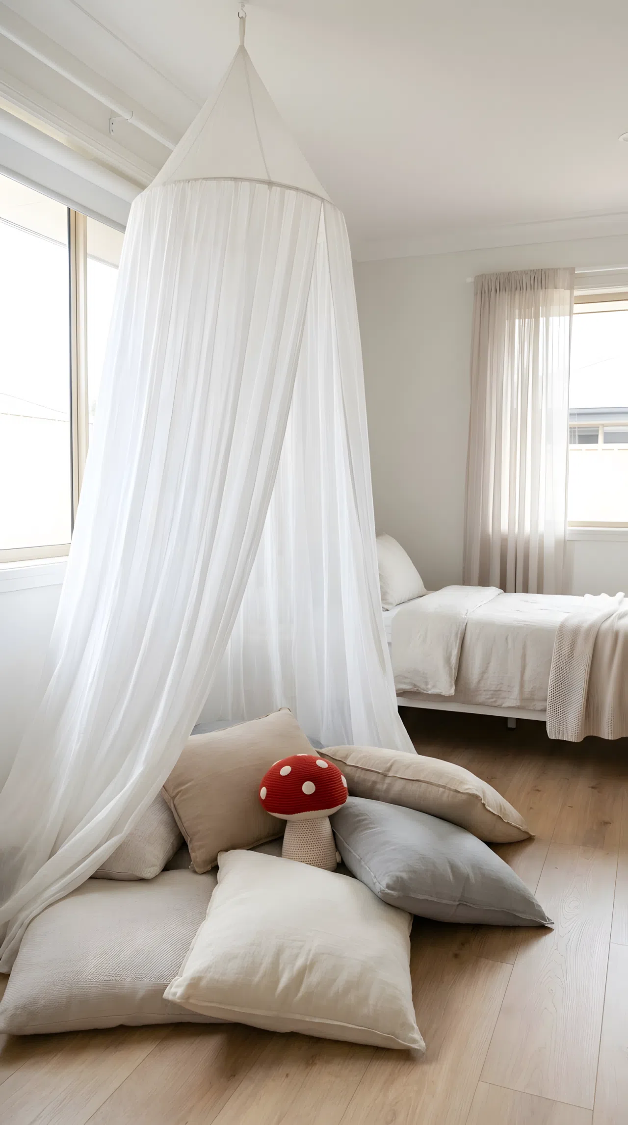 Real-life photo of a kids bedroom corner with a soft white fabric canopy tent draped over floor cushions, small red mushroom-shaped plush toy sitting among the cushions, neatly made bed visible in the background, bright natural light, beautifully arranged, organized, clean, no people, no text overlays