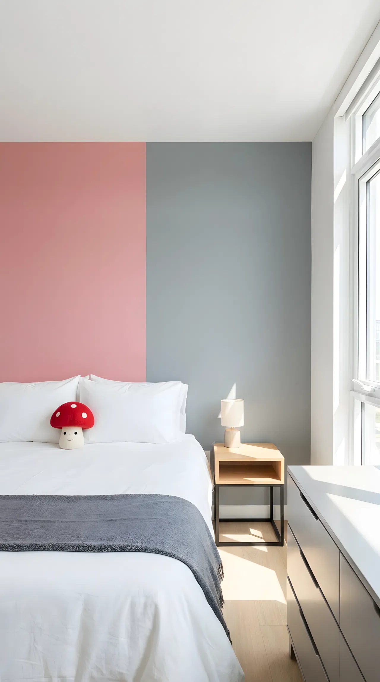 Real-life stock photo style bedroom, neatly made bed against a color block wall split between pink (RGB 226,150,160) and grey, streamlined modern furniture, small red mushroom shaped toy on the pillow, beautifully arranged, organized, clean, bright natural light, vibrant colors, no people, no text overlay