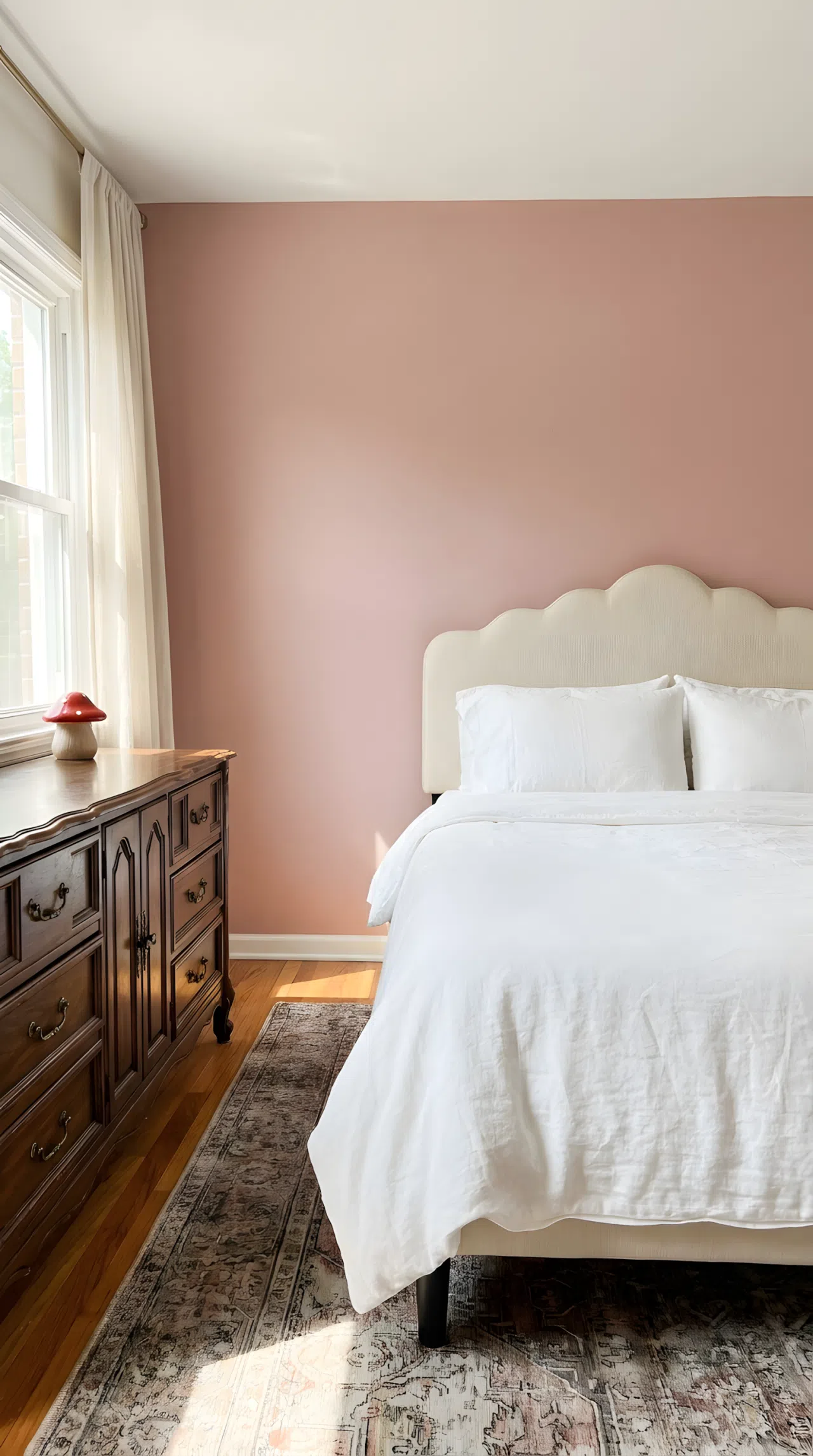 Real-life stock photo style bedroom, neatly made bed with a scalloped headboard against a muted pink wall, carved vintage wood dresser nearby, small red mushroom shaped toy sitting on the dresser, beautifully arranged, organized, clean, bright natural light, vibrant colors, no people, no text overlay