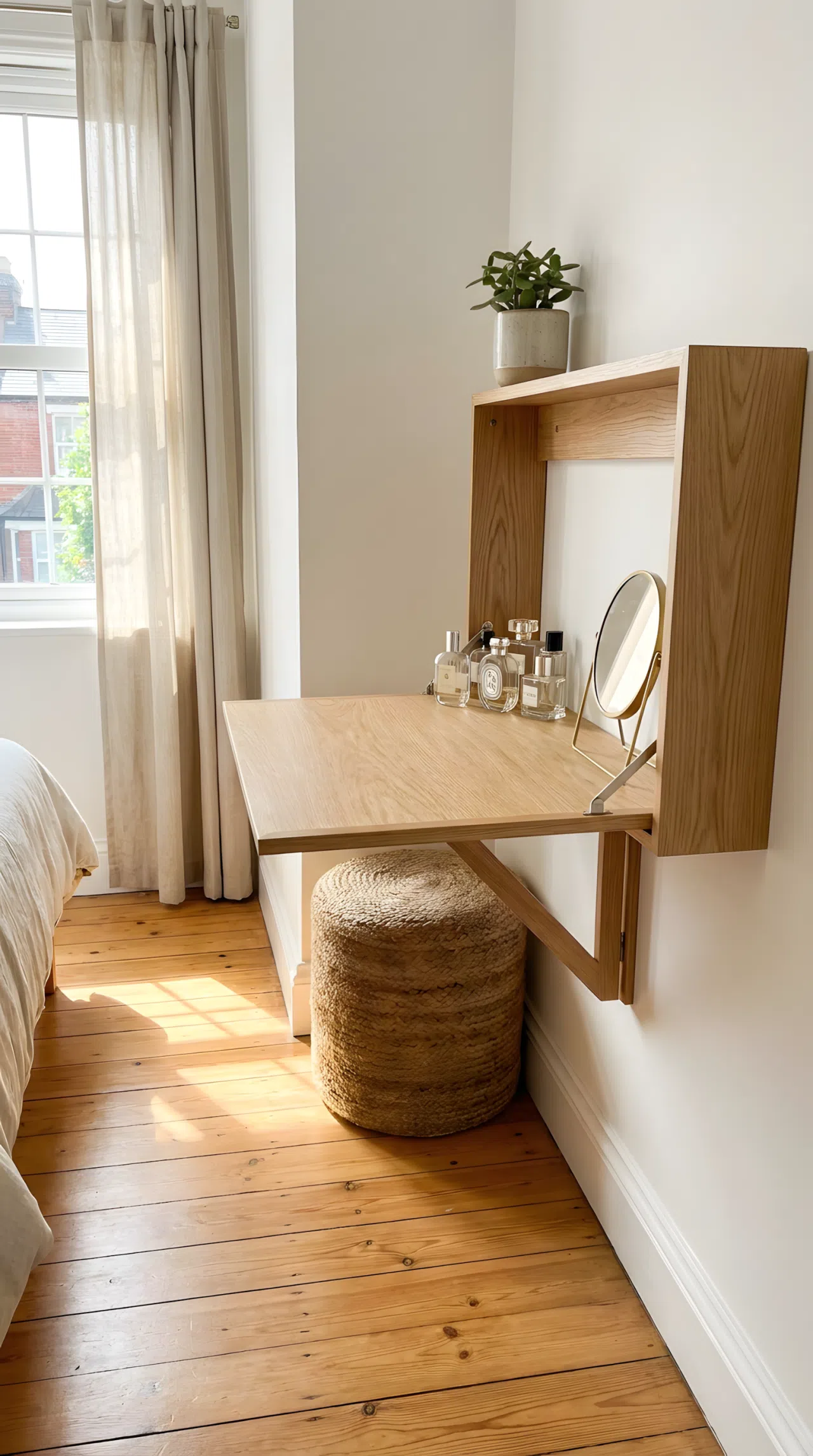 Stock photography, real-life practical small bedroom setup, a sleek wall-mounted folding oak desk unfolded in a compact bedroom space, neatly made surface with organized glass perfume bottles and a small brass mirror, a woven stool pushed neatly underneath, clean wooden floor in a narrow walkway, beautifully arranged room corner, bright natural light streaming from nearby window.