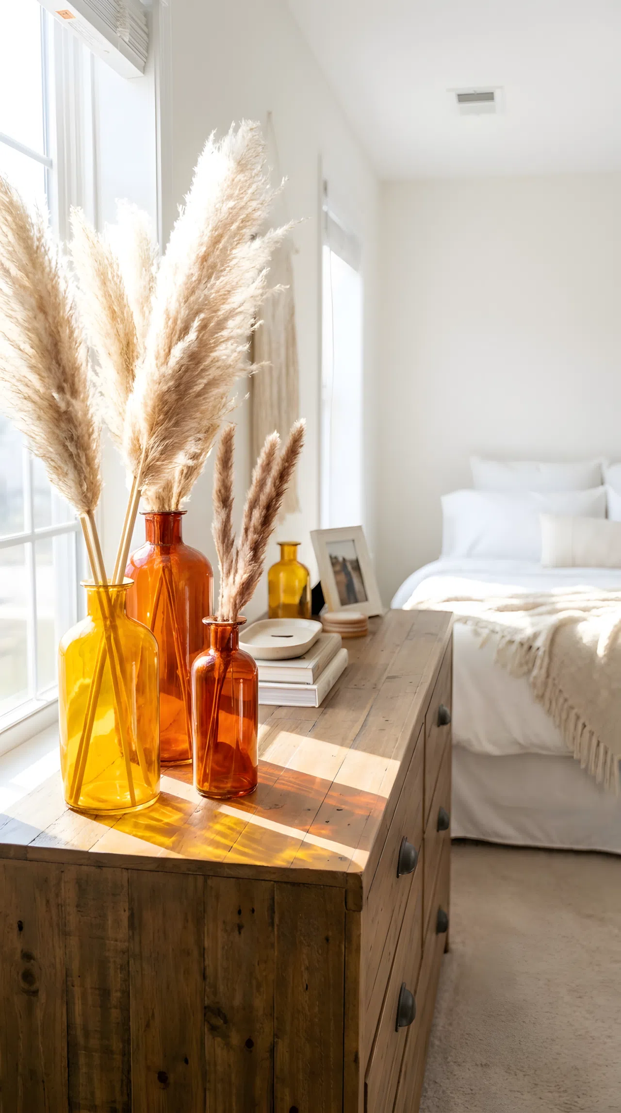 Real-life stock photo of a wooden bedroom dresser adorned with amber glass RGB(255, 191, 0) and burnt orange glass RGB(204, 85, 0) vases holding delicate dried pampas grass. The background shows a neatly made bed. Beautifully arranged, organized, clean, bright natural light reflecting through colored glass, vibrant rich tones, realistic home setup.
