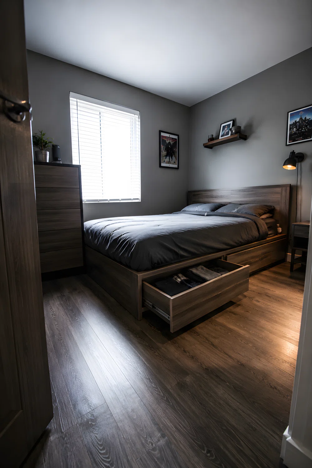 Wide-angle photograph of a compact real bedroom, low platform storage bed with one drawer pulled open showing folded clothes, grey bedding, minimal decor, bright window light from the side, dramatic angle showing full room depth, natural vibrant tones, realistic photography not 3D render.