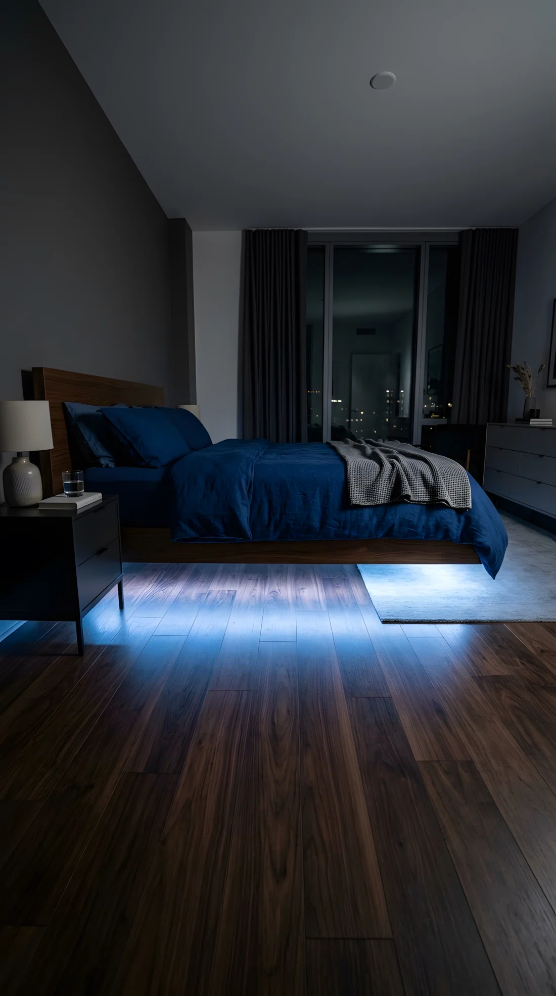 A wide-angle photograph of a modern bedroom at night, soft cool-white LED strip lighting (RGB 173,216,255) glowing beneath a floating bed frame casting a gentle halo onto the dark wood floor, the rest of the room dim, vibrant navy bedding above, dramatic low camera angle from floor level capturing the full underglow effect, photographic stock-image realism, clean and organized room.