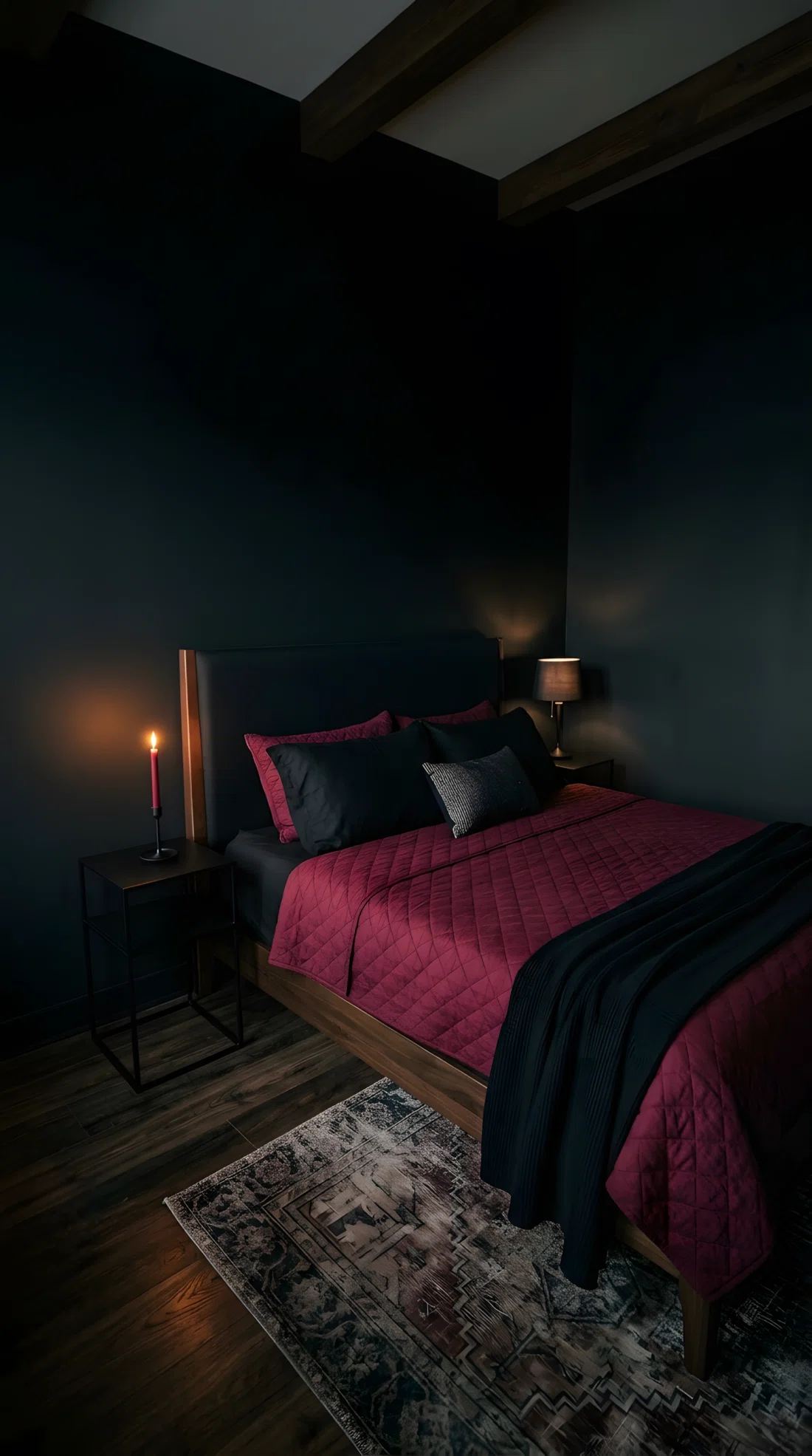 Wide-angle real photo of a black bedroom with a burgundy quilt layered under black pillows on the bed, single red taper candle on a black nightstand, small table lamp positioned low and close to the bed casting warm light across the headboard fabric, matte black walls, dramatic wide camera angle showing full room depth, photographic stock photo style, vibrant deep red tones, quilt color RGB 122,20,25