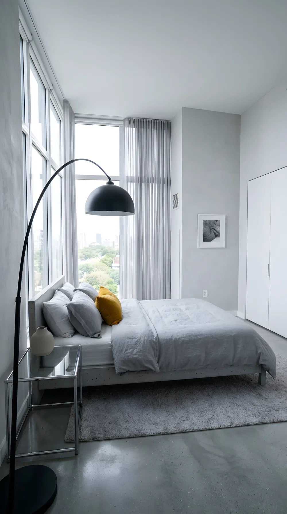 A wide-angle real-life photo of a sleek modern bedroom with a low platform bed, glass accent nightstand, floor to ceiling white curtains, monochrome gray and white color scheme with one bold accent color pillow, single statement floor lamp, neatly made bed, bright natural light, clean minimalist organized space, photographic stock photo style, dramatic wide camera angle.