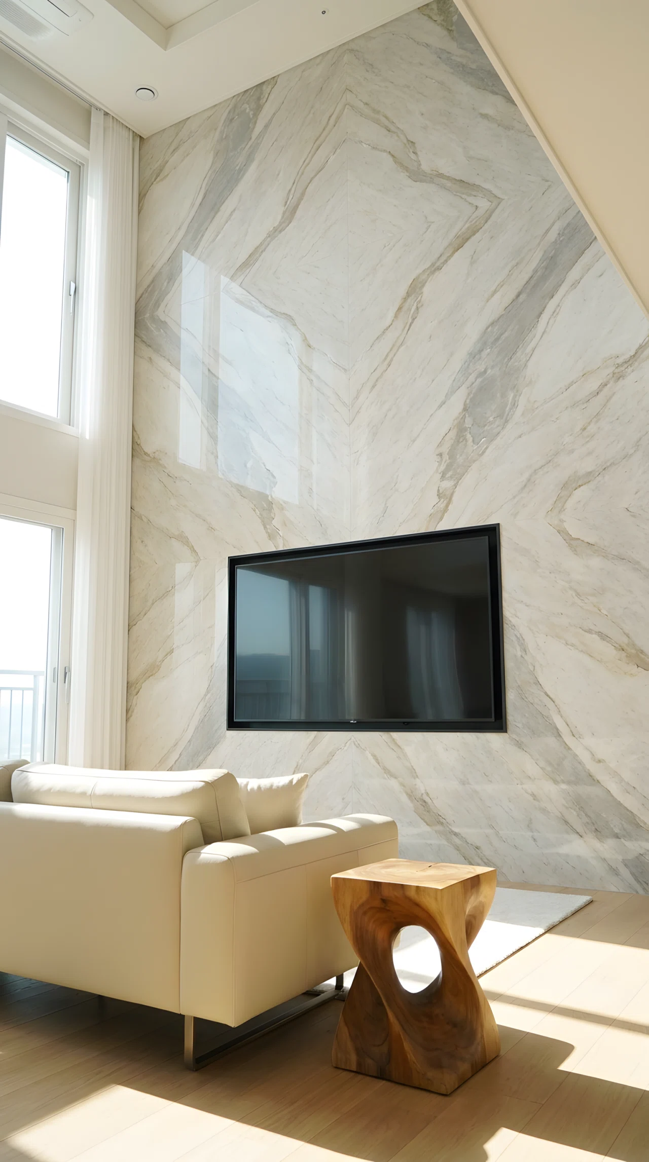 Wide-angle real-life photograph of a living room with a large book-matched marble slab TV wall in soft grey and warm beige veining (base RGB 232,227,219, vein RGB 138,128,112), a flatscreen TV mounted flush with a thin matte black frame around it, minimal furniture including a low cream sofa and a single sculptural side table, dramatic low wide camera angle capturing the full marble pattern from floor to ceiling, bright natural daylight streaming in from a side window to highlight the stone veining, no people in the frame, vibrant true-to-life color rendering, realistic stock photography, not a 3D render or illustration.