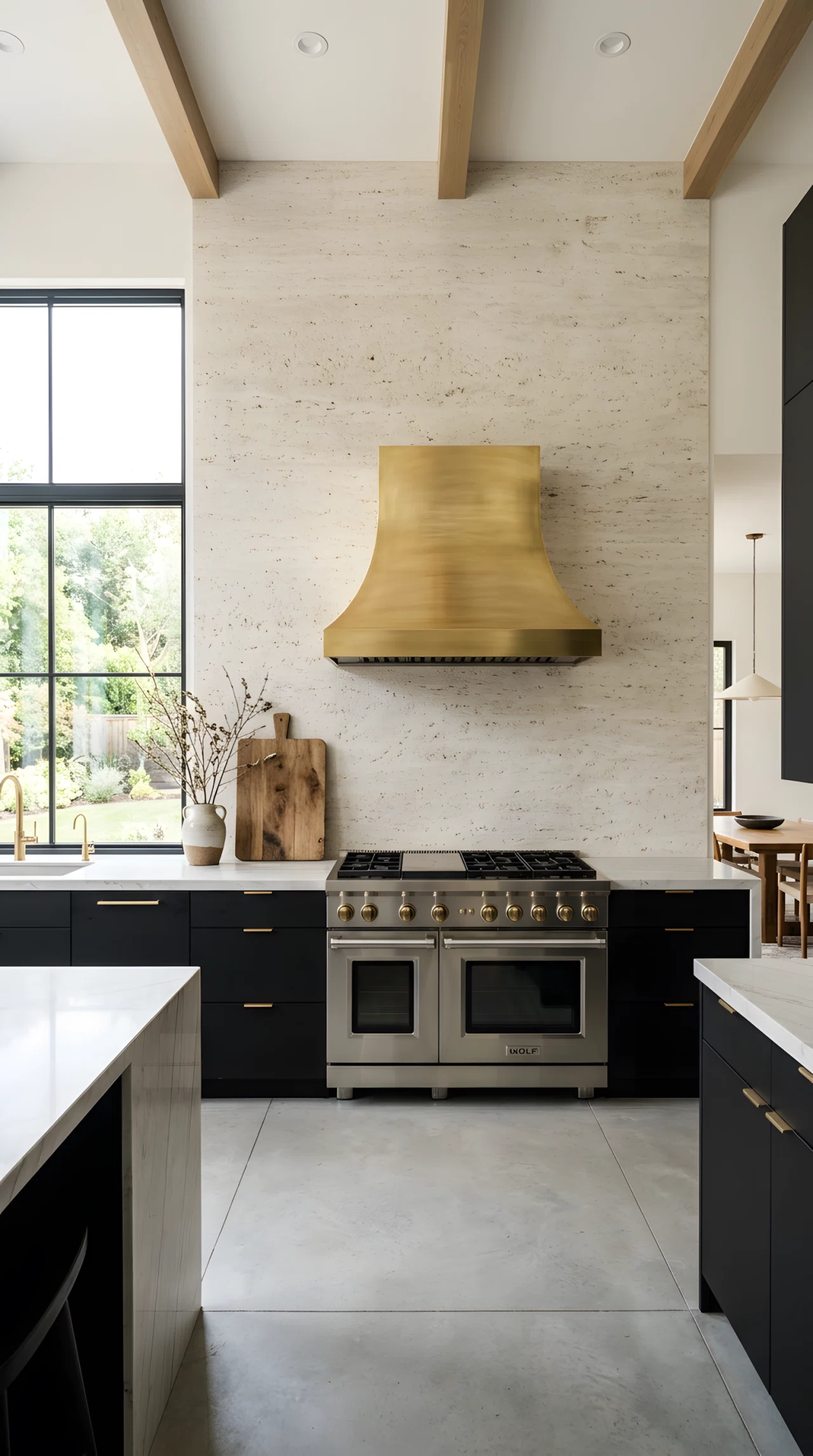 Wide-angle real-life photograph of a modern luxury kitchen with a floor-to-ceiling textured stone backsplash in soft travertine tones behind a professional-style range, brushed brass range hood, matte black lower cabinets, minimal countertop clutter with only a wooden cutting board and a ceramic vase, bright natural light entering from a side window, dramatic wide camera angle showing the full statement wall, realistic stock-photography style, no people.