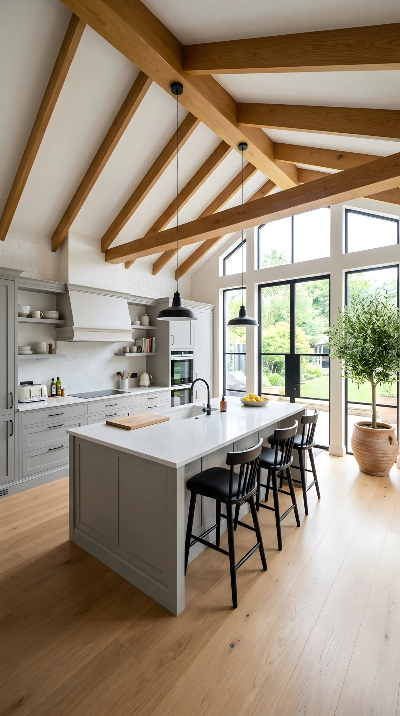 Wide-angle real-life photograph of a modern luxury kitchen with exposed warm wood ceiling beams running across a white ceiling, two simple black pendant lights hanging between the beams above an island, bright natural light entering from tall windows, matte cabinetry below in soft grey, vibrant potted olive tree in the corner as an accent, dramatic wide camera angle capturing the full ceiling structure and room below, realistic stock-photography style, no people.